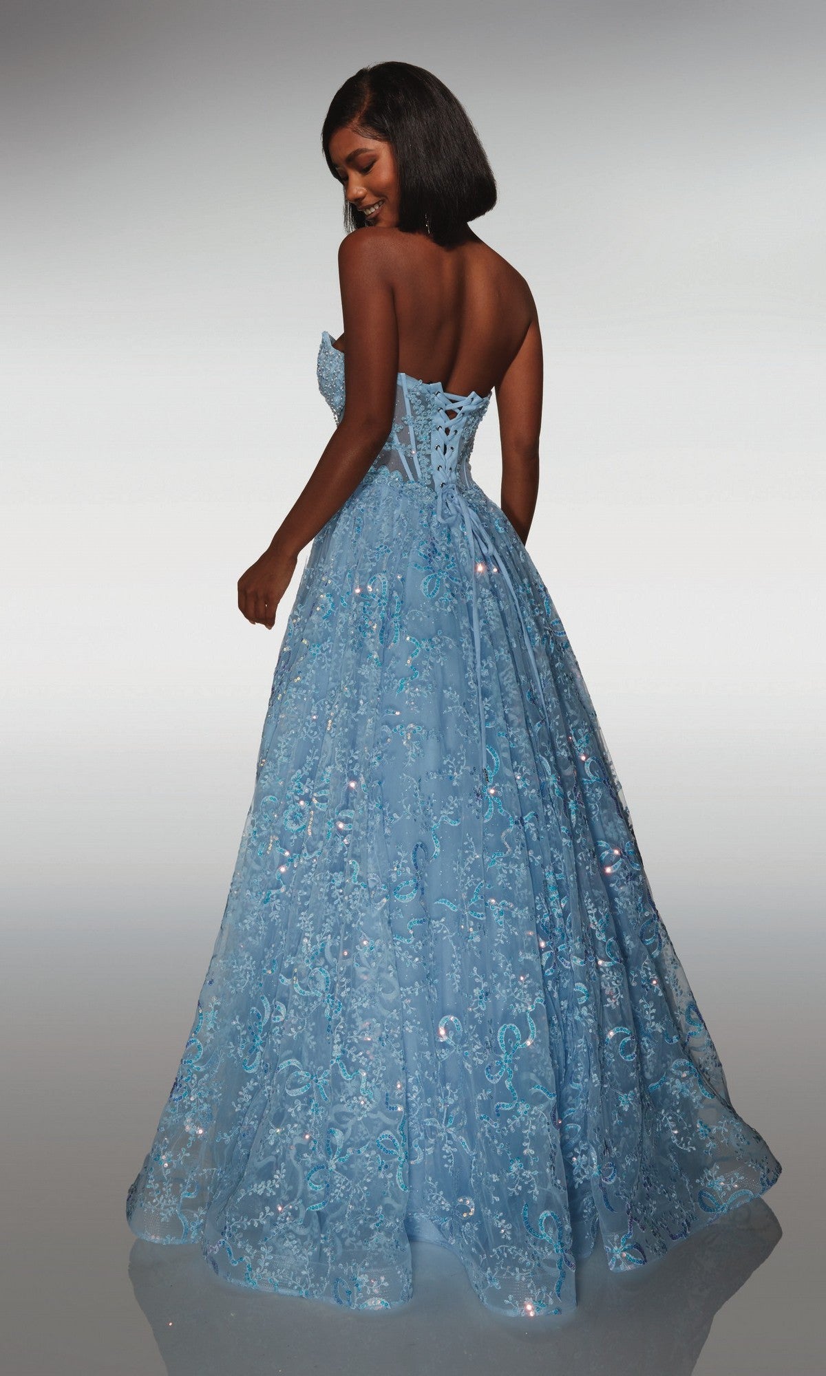 A woman stands with her back to the camera wearing the Alyce 62033 long formal dress, featuring a strapless beaded corset bodice, floor-length light blue skirt, and sequin lace details.